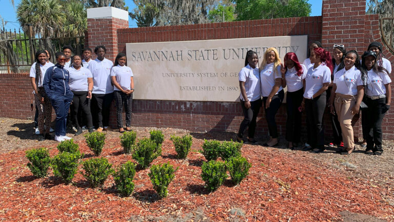 A group of teenage girls stands in front of the Savannah State College entrance sign.