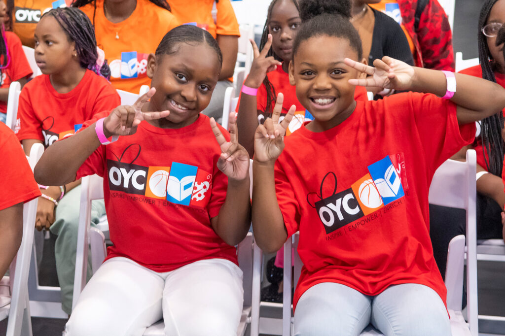 two young girls wearing OYC Miami shirts flash the peace sign while seated on a bleacher