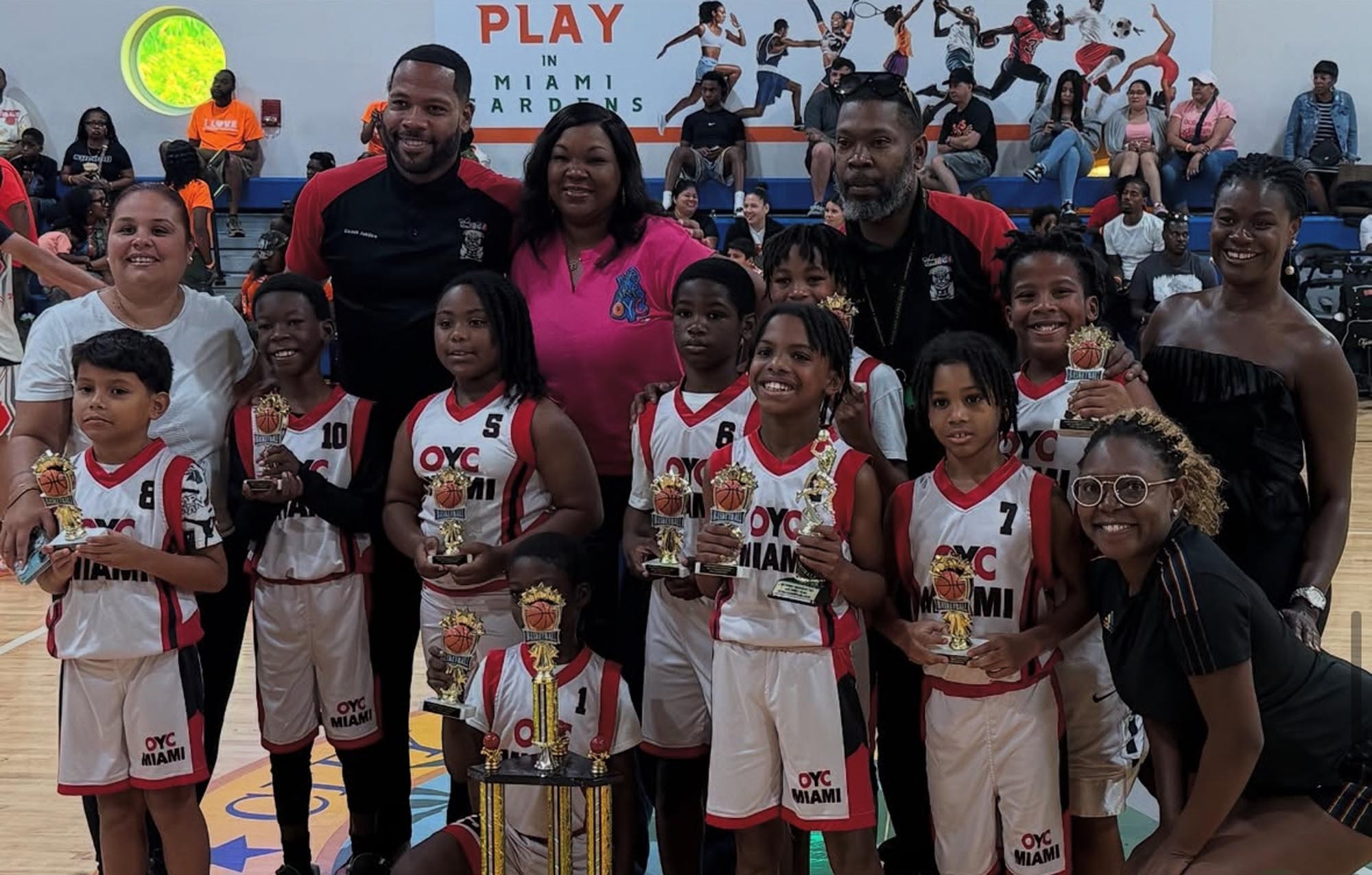 A group of young basketball players holding trophies stand alongside their coaches and parents