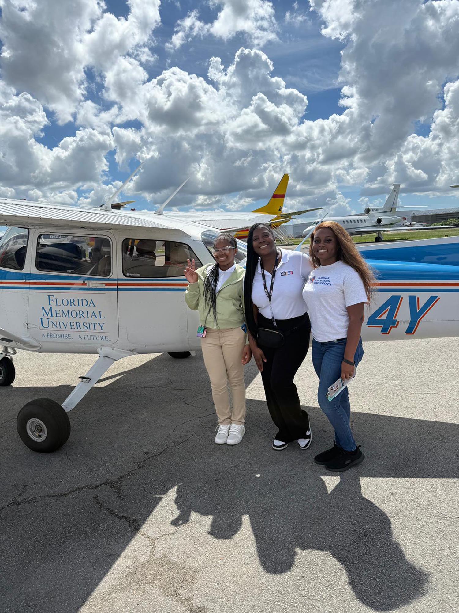 three young ladies pose for a photo next to a small passenger plane on a tarmac