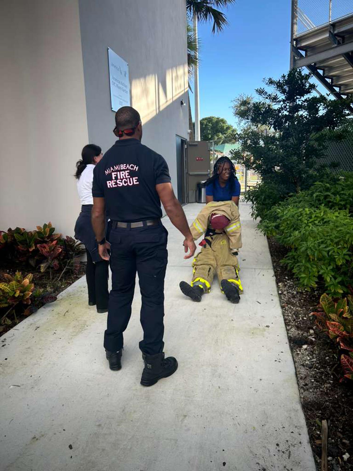 a young man pulls a dummy across a sidewalk as firefighter instructors look on