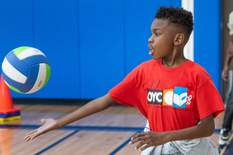 a boy wearing a red OYC Miami shirt hits a volleyball in a gym