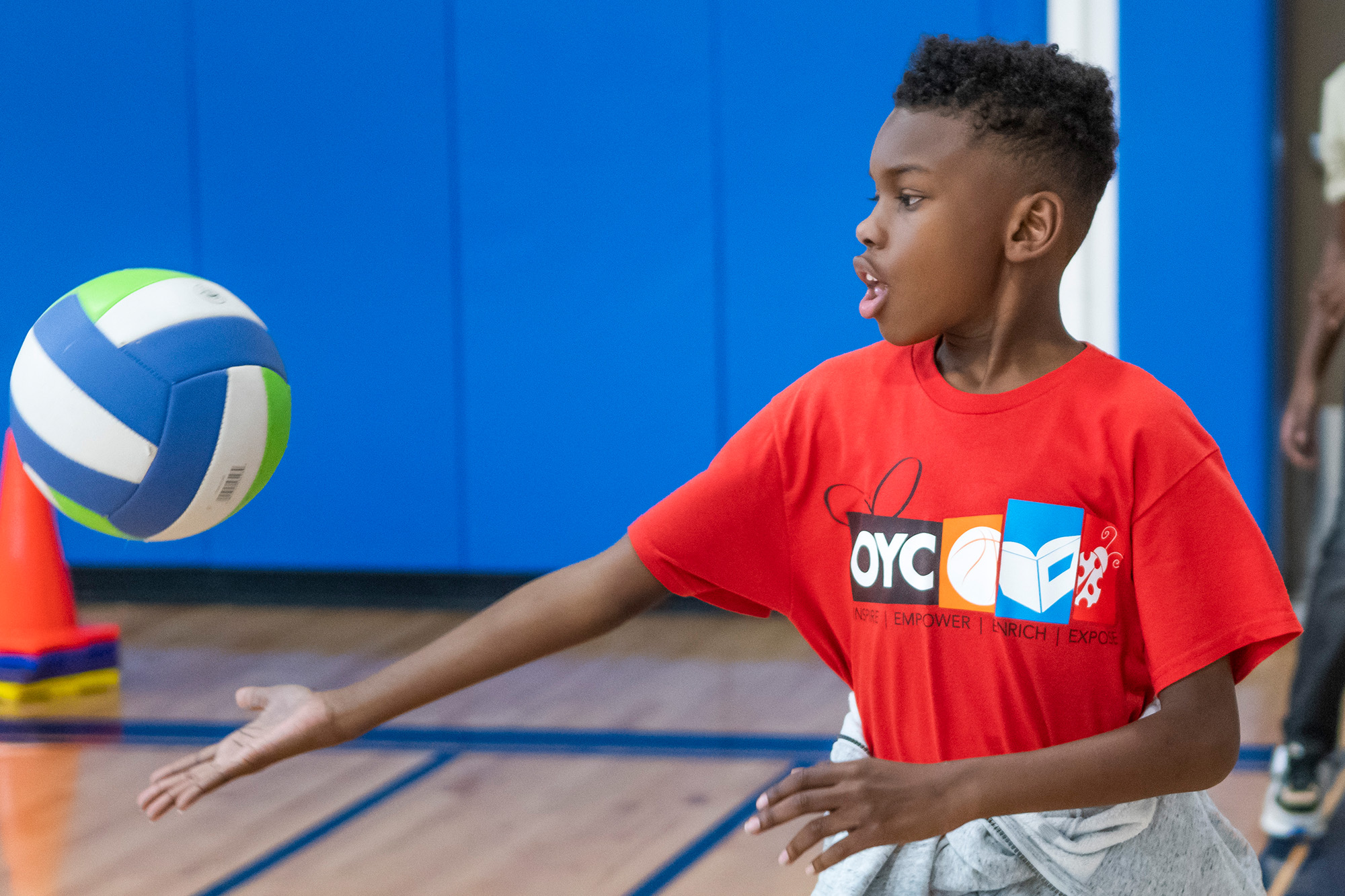 a boy wearing a red OYC Miami shirt hits a volleyball in a gym