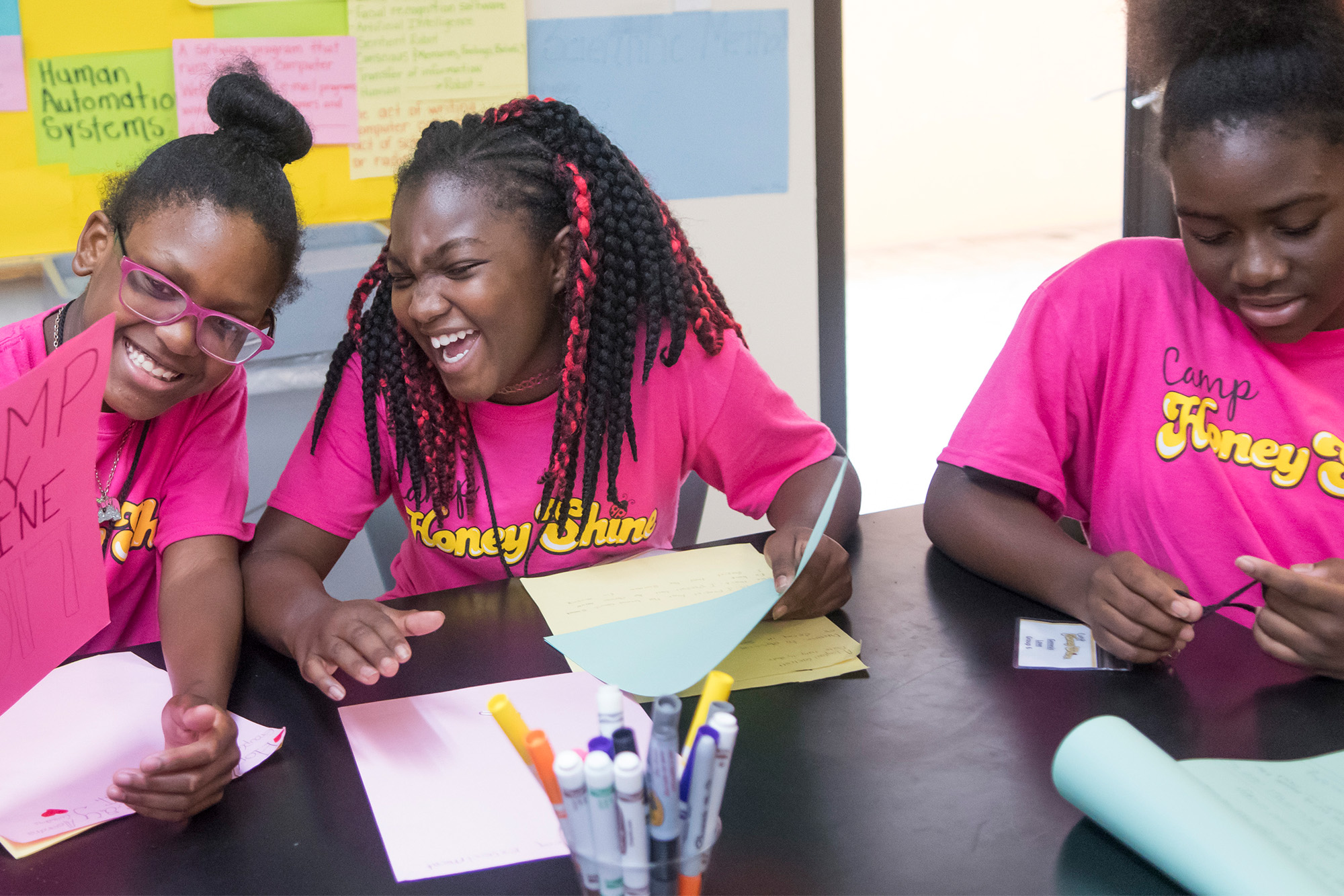 three teen girls wearing pink tees sit and do arts and crafts