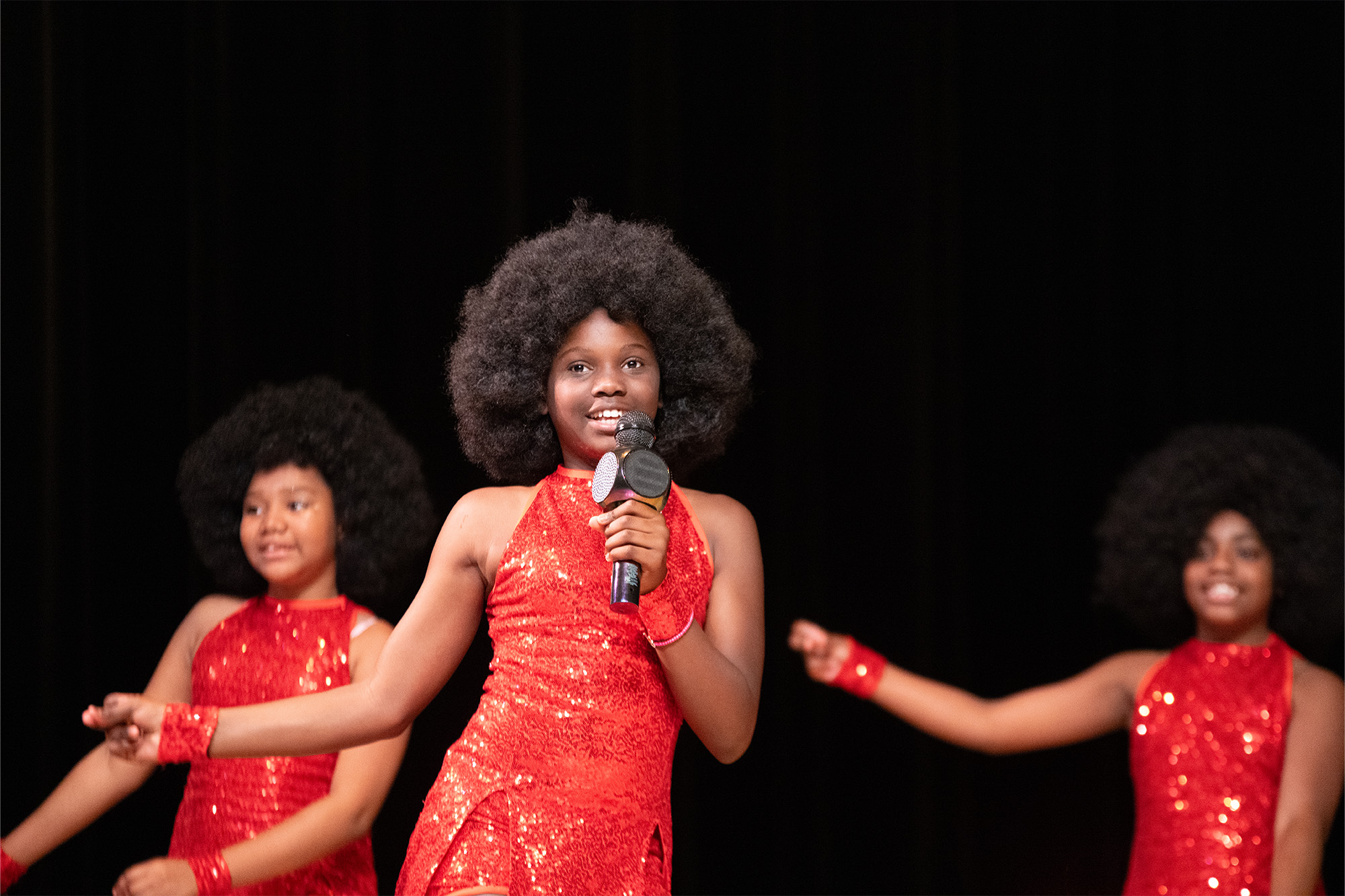 three girls wearing red dresses perform on stage at a showcase